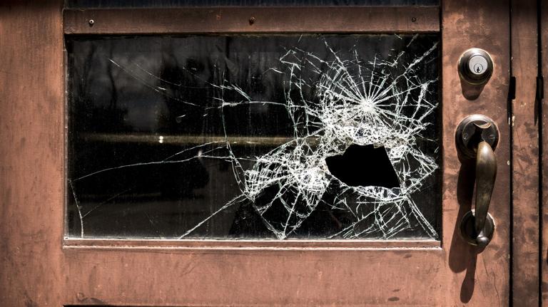 A door with a glass window features a large, shattered area with spiderweb cracks around a hole. The metal handle and lock are visible, conveying neglect.