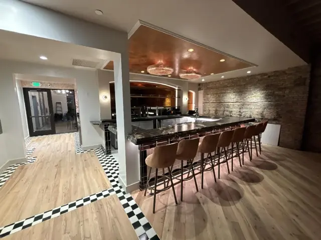 Vagabondo's finished bar in Excelsior with a dark stone counter, leather stools, a copper-leaf ceiling panel, and the building's original brick wall cleaned and uplit