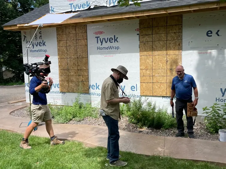 Crew filming outside a house wrapped in housewrap partway through reconstruction