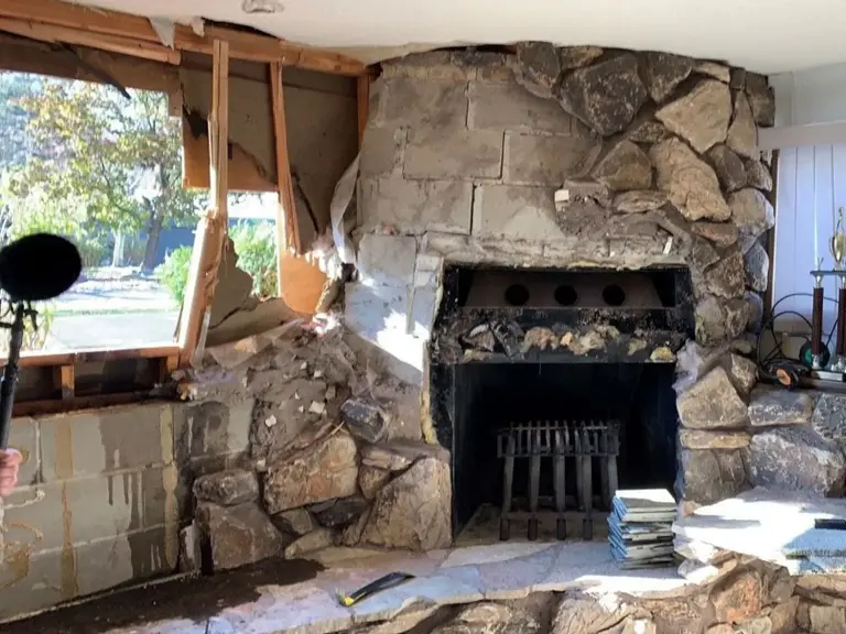 Living room wall broken open beside a floor-to-ceiling fieldstone fireplace, with snapped framing, torn drywall, and daylight through the opening after a vehicle struck the house