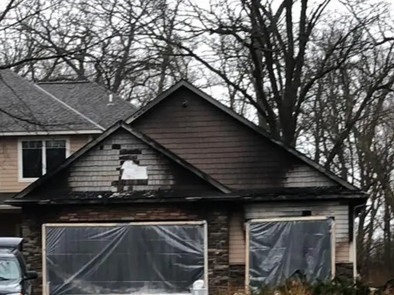Fire-damaged garage gable with burned cedar shakes, charred soffit and fascia, and both overhead door openings sheeted off