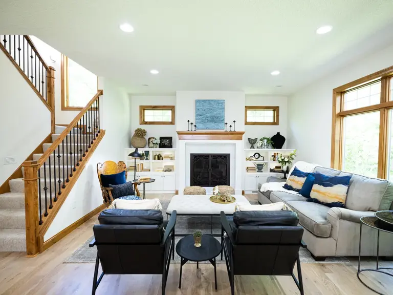 The same living room rebuilt with a white fireplace surround, white built-in shelving, and an open staircase with black metal balusters