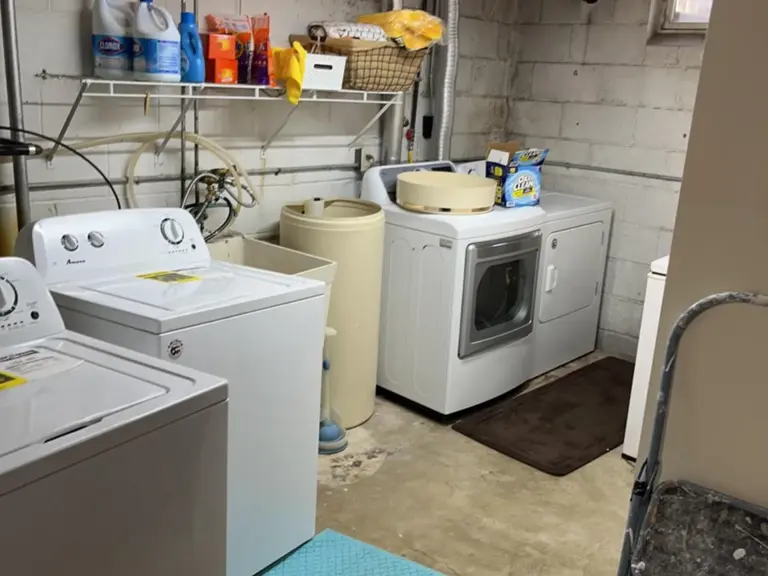 Original unfinished laundry area with an exposed block wall, bare concrete floor, and open piping before restoration