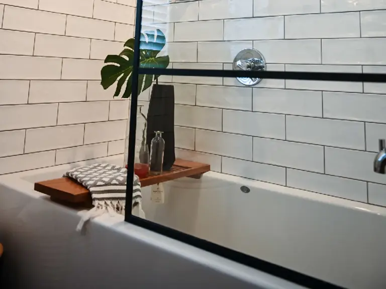 The same tub alcove rebuilt with white subway tile, a black grid glass panel, and a teak bath shelf