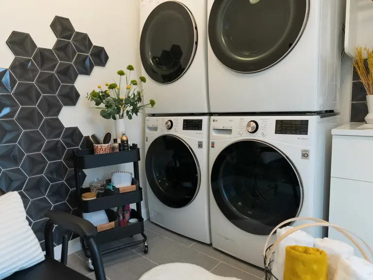 The same stacked washers and dryers after restoration, set against a black geometric-tile wall beside the laundry-sink cabinet