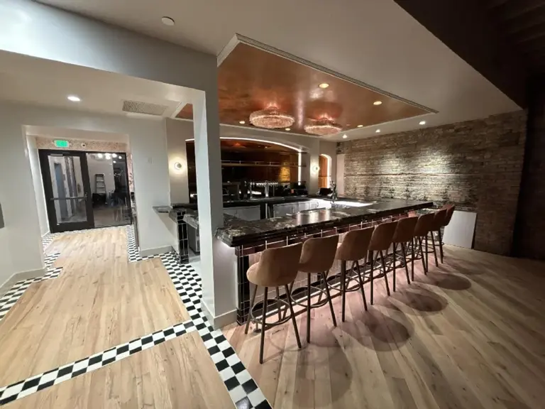 The same corner rebuilt as the new bar, with a dark stone counter, leather stools, a copper-leaf ceiling panel, and the original brick wall cleaned and uplit