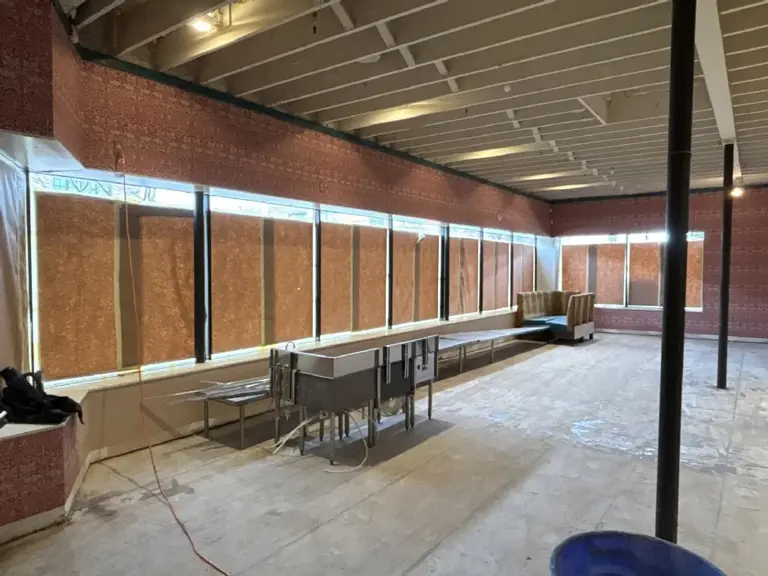 Restaurant dining room stripped to bare concrete during renovation, with the storefront windows boarded over, patterned wallpaper above them, and the original ceiling joists exposed