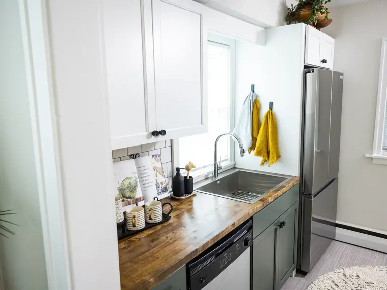 Renovated kitchen sink wall with butcher block counter, white upper cabinets, green lower cabinets, and subway tile
