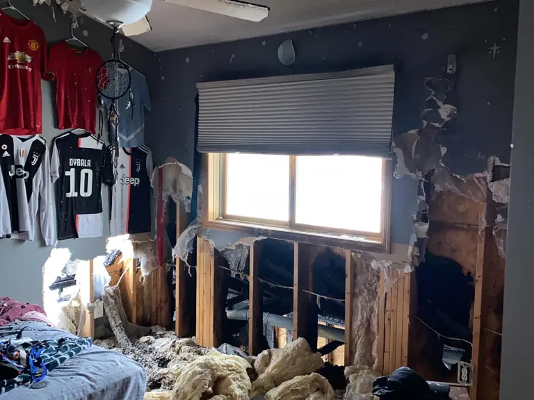 Upstairs bedroom wall opened to charred studs and wet insulation after smoke and fire damage from a garage fire