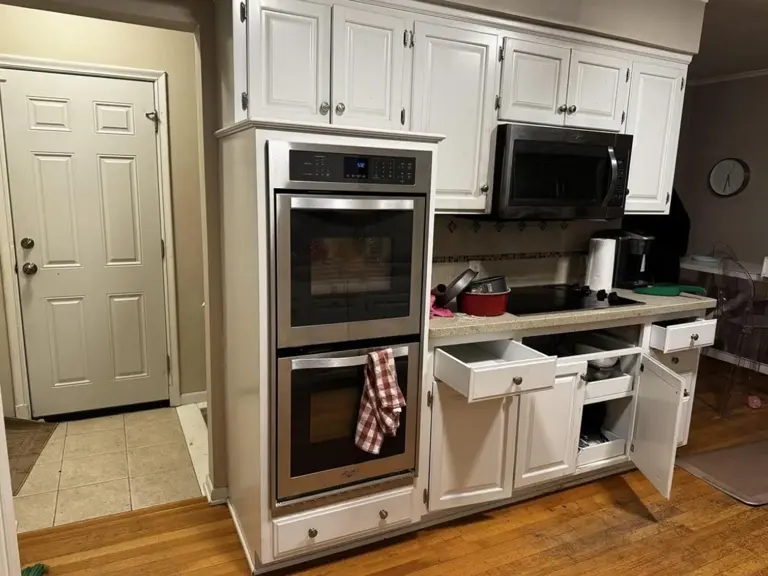 Original kitchen cooking wall before fire restoration, with white raised-panel cabinets, stacked wall ovens, a black cooktop and an over-range microwave beside the entry door