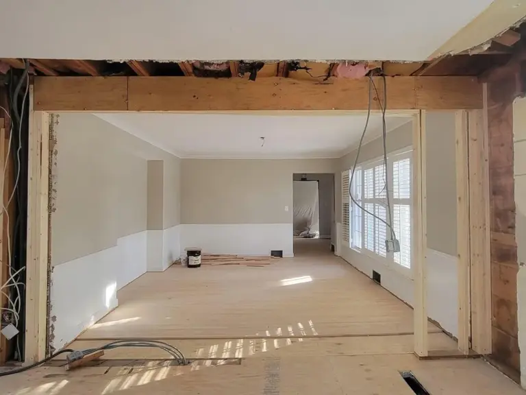 Kitchen stripped to studs and subfloor after fire damage, with a new structural header framing the opening into the dining room and charred joists visible overhead