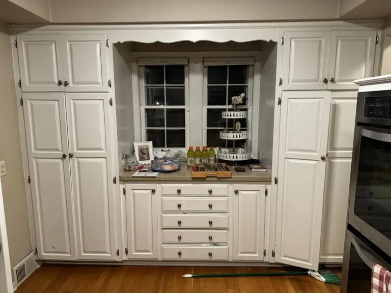 Kitchen window flanked by full-height raised-panel pantry cabinets, with a scalloped valance and a laminate counter beneath, before fire damage restoration