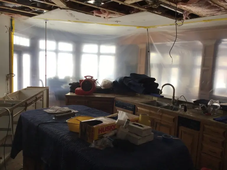 The kitchen under containment during water damage mitigation, with the ceiling opened, plastic sheeting hung, and cabinets covered