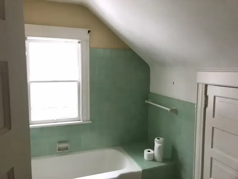 Dated upstairs bathroom before remodeling, with green wall tile and a tub under a sloped dormer ceiling beside a window
