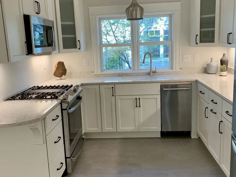 The same U-shaped kitchen rebuilt with white shaker cabinets, quartz counters, a stainless gas range, and tile flooring, the sink centered under the window