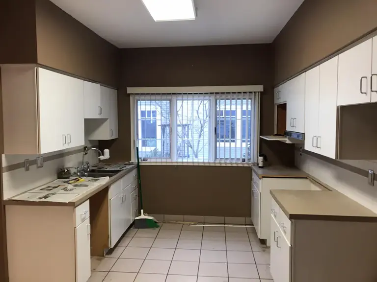 Dark Lake of the Isles kitchen before restoration, with white laminate cabinets, brown walls, white tile floor, and a window over the sink in a U-shaped layout