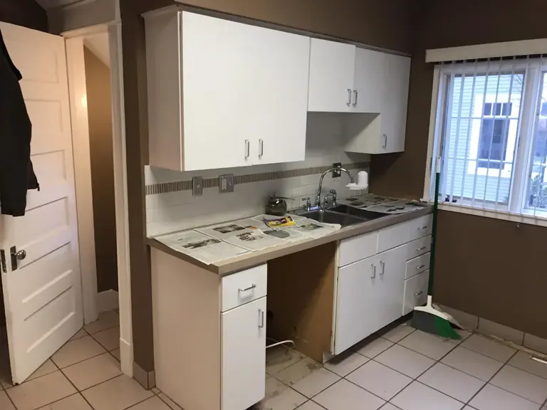 Original kitchen wall with a double sink, white laminate cabinets, and a window to the right before fire restoration