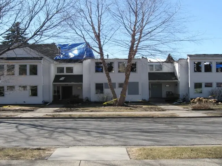 Fire-damaged row of townhomes with a tarped roof, boarded windows, and debris in the yard before restoration