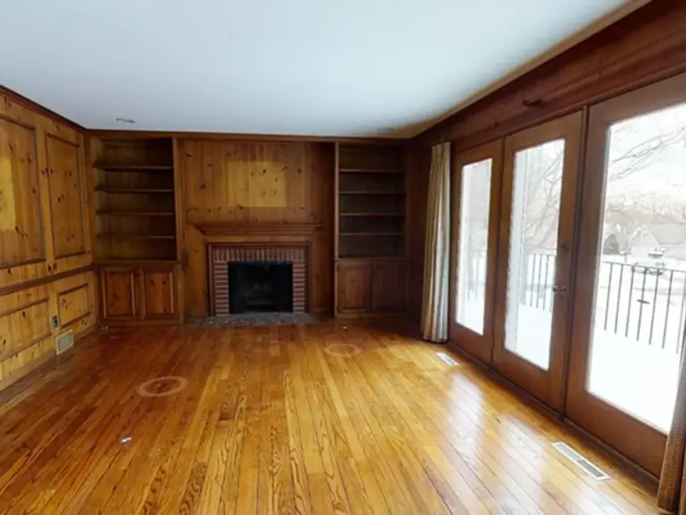 Dated wood-paneled den with a brick fireplace, built-in shelving, and French doors out to a snowy deck before ice dam restoration