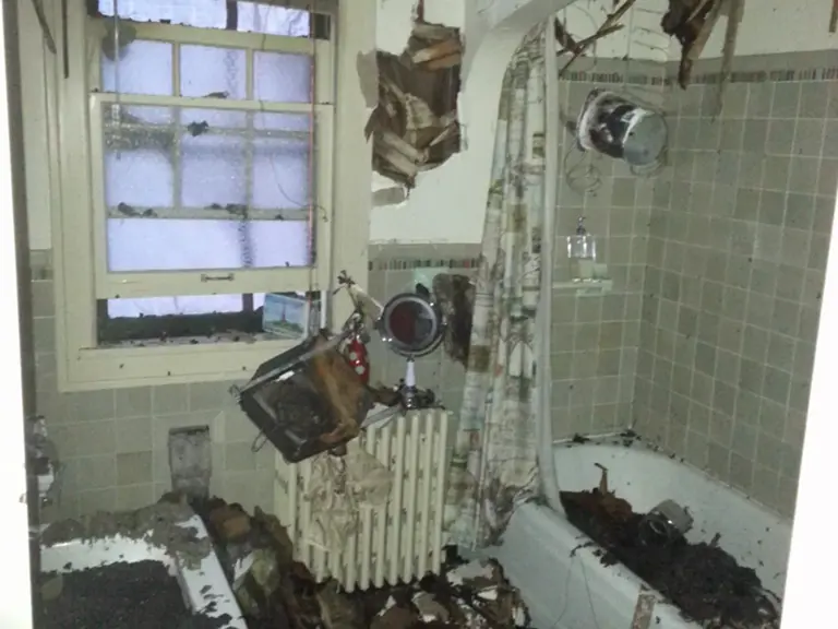 Fire-damaged bathroom with a soot-covered tub, radiator, and debris hanging from the opened ceiling