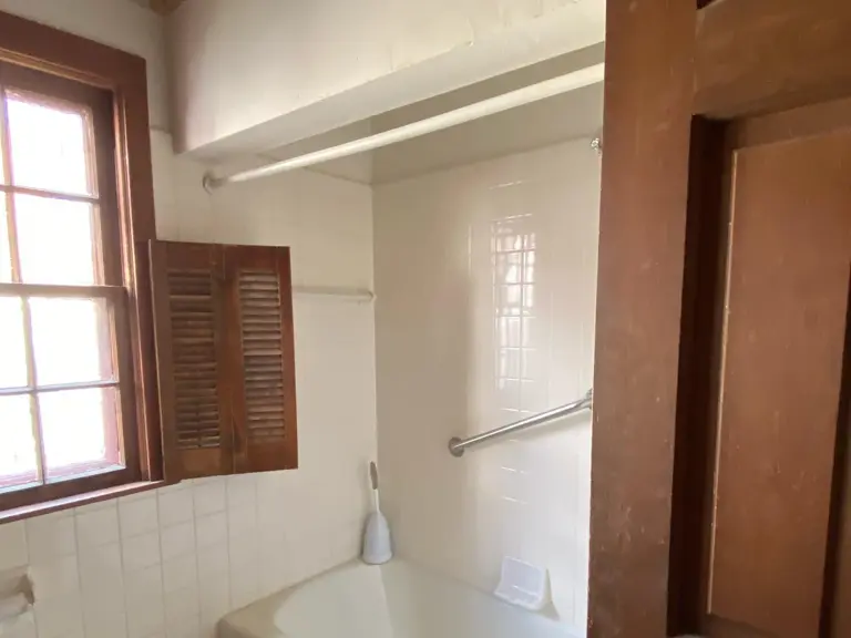 Original 1940s bathroom with a built-in tub, small white wall tile, a wood-shuttered window, and a stained-wood door before the remodel