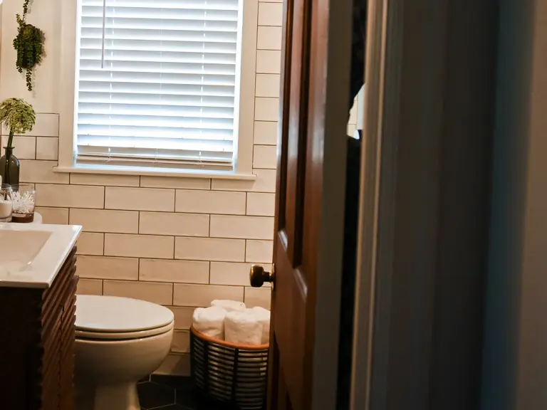 The same wall remodeled with a walnut slat-front vanity, white subway tile, a black faucet, white window blinds, and black hexagon floor tile