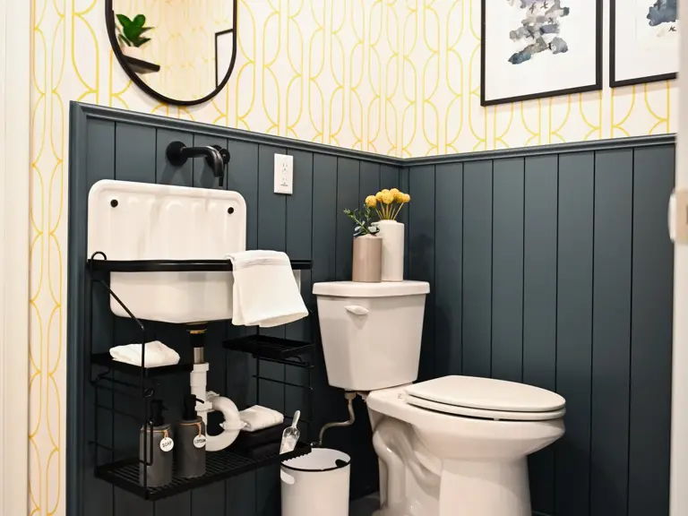 The same basement bathroom rebuilt with dark gray shiplap wainscot, yellow patterned wallpaper, an oval mirror, and a new wall-hung sink