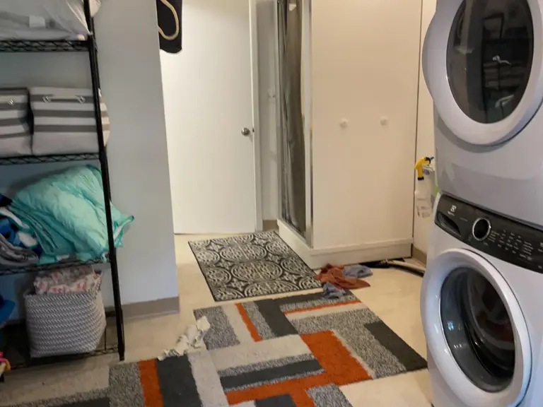 Basement laundry area before restoration, with a stacked washer and dryer, wire shelving, and layered rugs on the original flooring