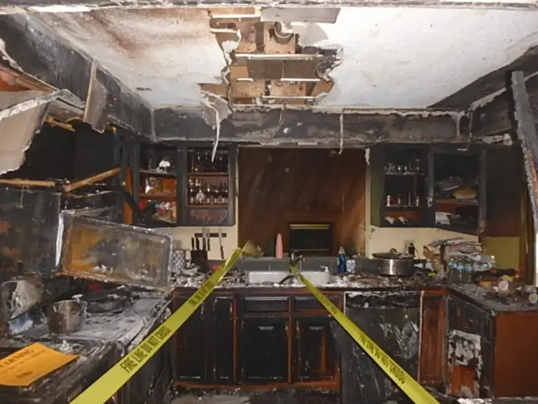 Fire-damaged kitchen behind fire line tape with charred cabinets and an opened ceiling before restoration