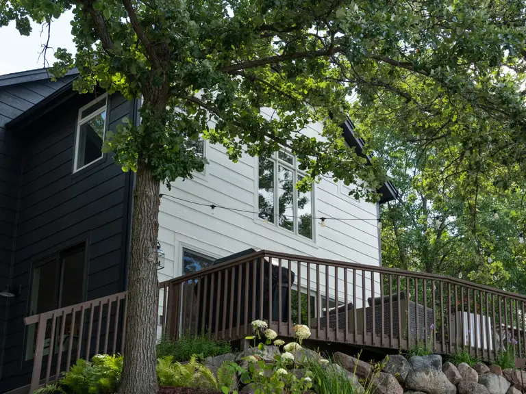 The same deck elevation repainted with white board-and-batten and charcoal siding above the boulder wall and mature oak