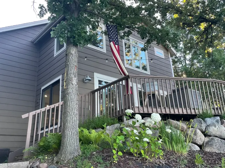 Grey-green lap siding and white trim on the lakeside deck elevation, above a boulder retaining wall, before the exterior refresh