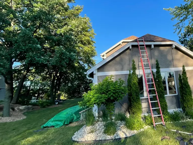 Taupe stucco gable mid-roof-replacement, with a ladder to the ridge and torn-off shingles scattered across the lawn after storm damage