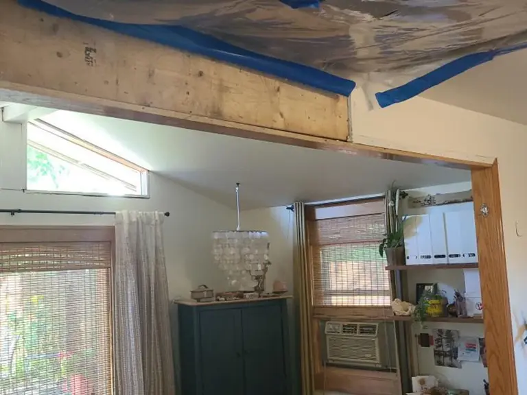 Living room stripped during storm and water damage repairs, with plastic containment taped across the ceiling and an exposed structural header over the opening