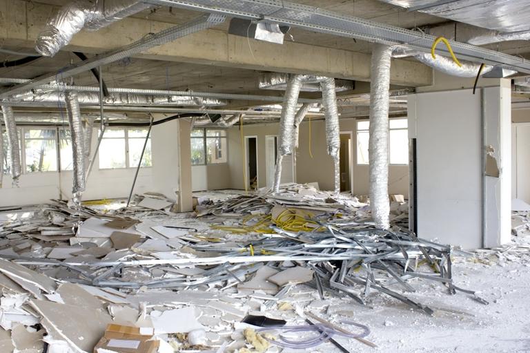 Empty office space under renovation with debris, broken ceiling panels, scattered wires, and exposed ducts. Daylight filters through the windows.
