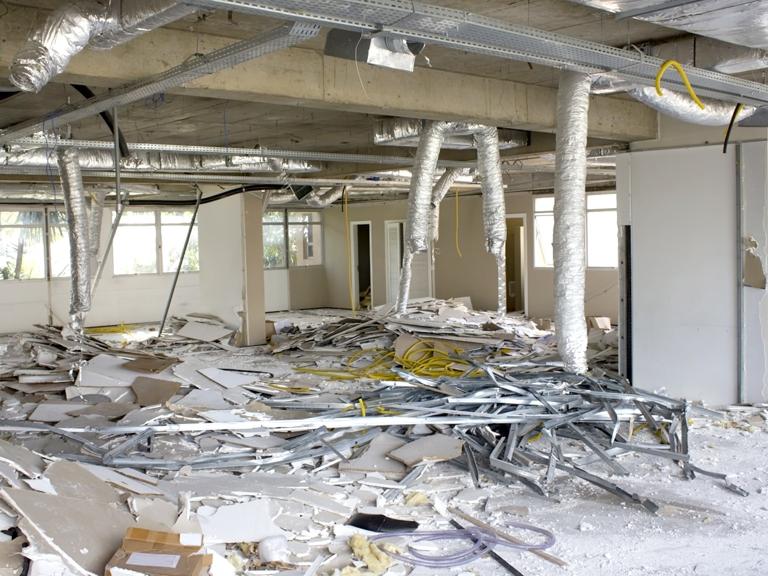 Empty office space under renovation with debris, broken ceiling panels, scattered wires, and exposed ducts. Daylight filters through the windows.