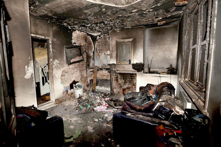 Charred room interior showing severe fire damage. The ceiling is blackened, and debris covers the floor. A burned sofa and broken window are visible, conveying destruction.