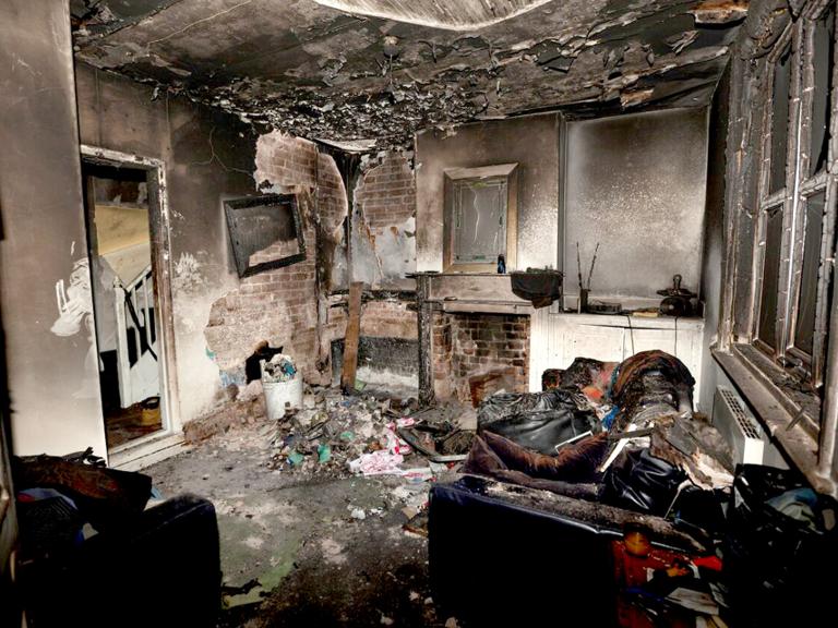 Charred room interior showing severe fire damage. The ceiling is blackened, and debris covers the floor. A burned sofa and broken window are visible, conveying destruction.