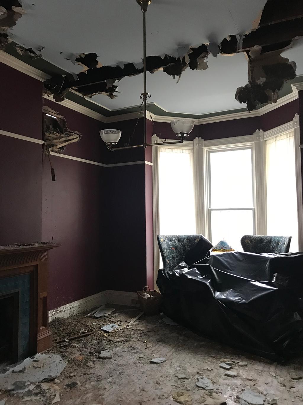 Damaged room with purple walls, debris-covered floor, and a large hole in the ceiling. Two armchairs by a bright window, evoking neglect.