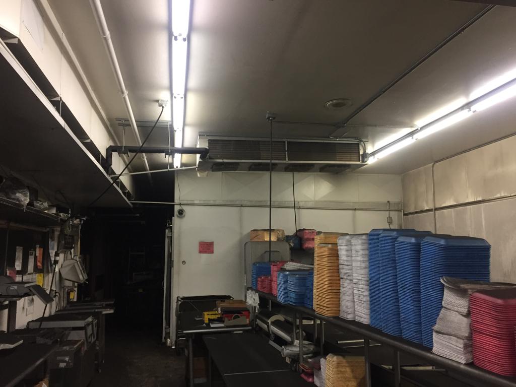 Industrial kitchen with overhead fluorescent lighting, featuring stacked colorful trays on metal tables. The room appears organized but dimly lit. Ungerman Construction, Prior Lake, MN