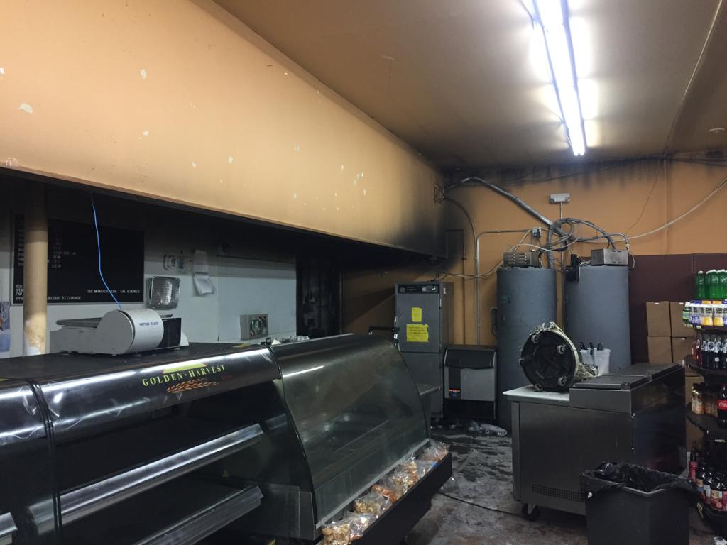 A dimly lit, abandoned kitchen with burnt walls and ceiling, containing empty display cases, industrial equipment, and shelves with beverages. The atmosphere is neglected. Ungerman Construction, Prior Lake, MN
