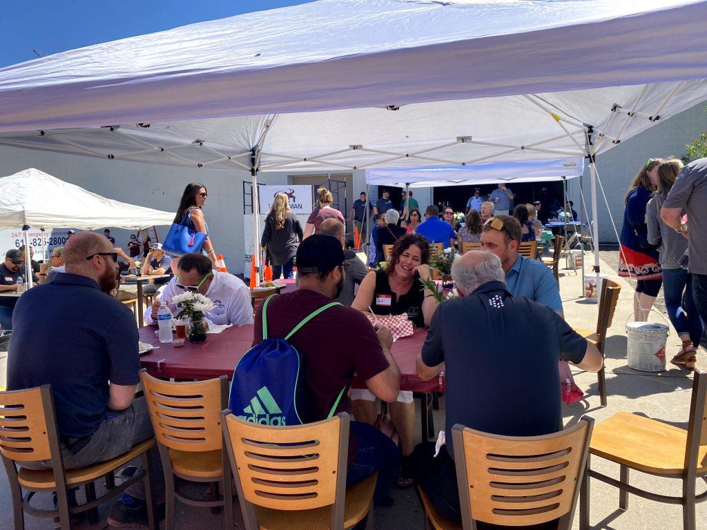 Outdoor gathering under white tents with people sitting at tables, engaged in conversation. The atmosphere is lively and social, with a clear blue sky.