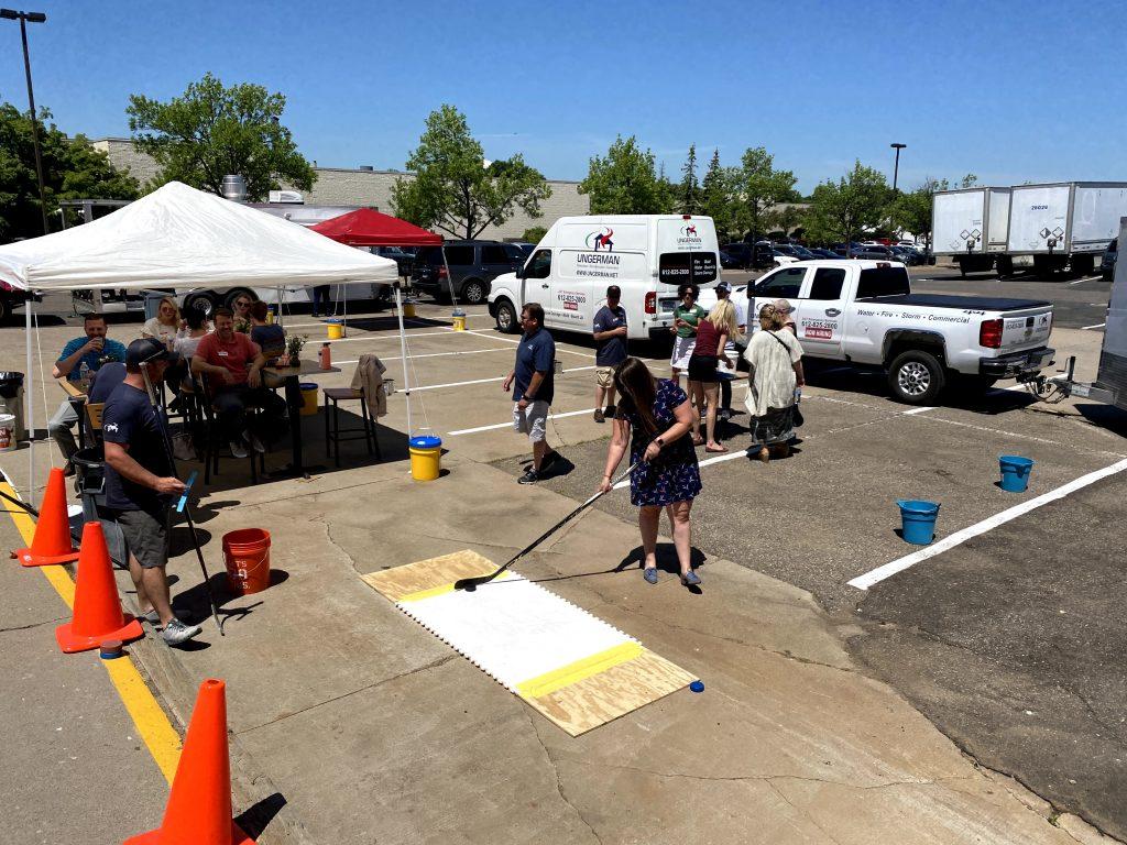 A woman plays street hockey on a makeshift rink in a sunny parking lot, surrounded by onlookers under a canopy. The atmosphere is lively and communal.