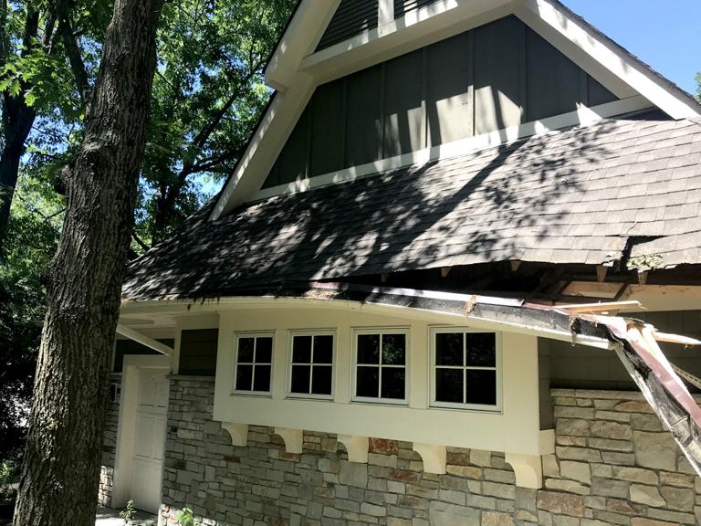 A house with a stone facade and shingle roof, damaged by a fallen tree branch. Sunlight filters through leaves, casting shadows on the roof.