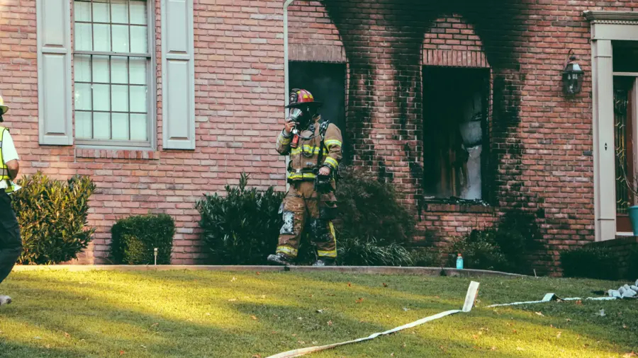 Firefighter in full gear stands beside a brick house with charred windows, indicating a recent fire. Sunlight casts long shadows on the grassy lawn.