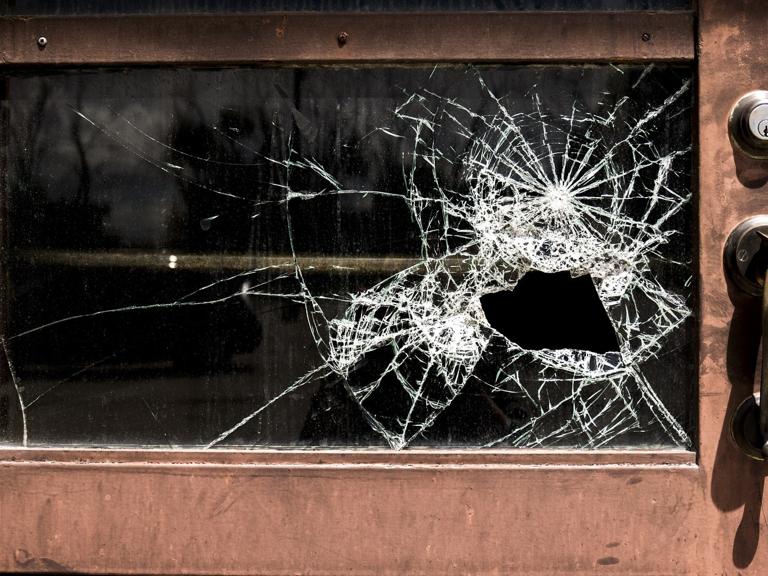 A door with a glass window features a large, shattered area with spiderweb cracks around a hole. The metal handle and lock are visible, conveying neglect.