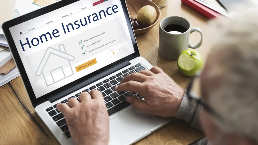 A person using a laptop displaying a home insurance website. Nearby are a cup of coffee, a green apple, and a bowl of fruit on a wooden table. The scene is calm and focused.