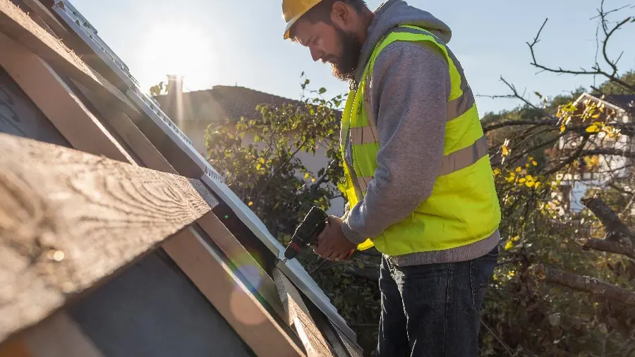 A construction worker in a yellow hard hat and vest uses a drill on a wooden roof. The sun shines brightly in the background, highlighting foliage. Ungerman Construction, Prior Lake, MN.