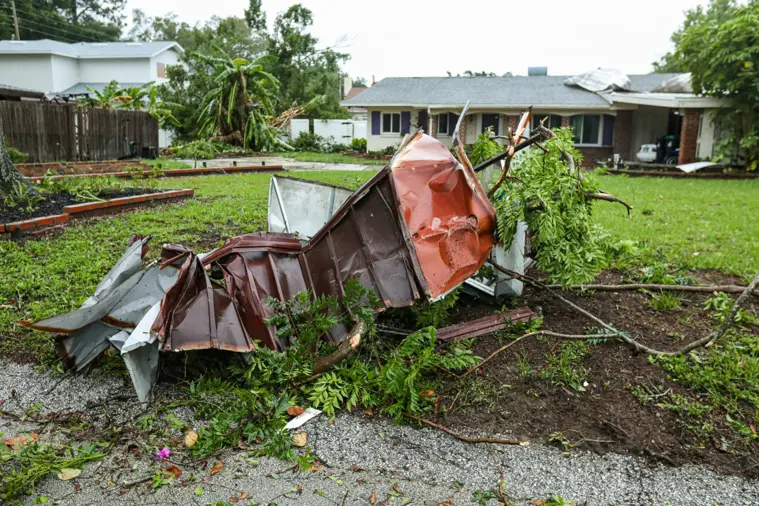 Twisted metal debris lies on a lawn in front of a house, amidst uprooted trees and scattered branches. The sky is overcast, suggesting storm aftermath. Ungerman Construction, Prior Lake, MN