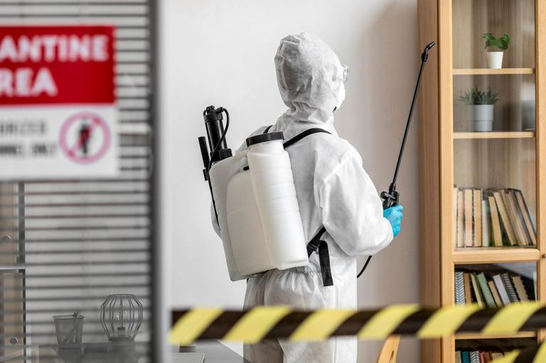 Person in protective gear sprays disinfectant in a quarantined room with Quarantine Area sign. Bookshelf and plants visible; caution tape in foreground.