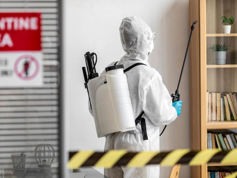 Person in protective gear sprays disinfectant in a quarantined room with Quarantine Area sign. Bookshelf and plants visible; caution tape in foreground.